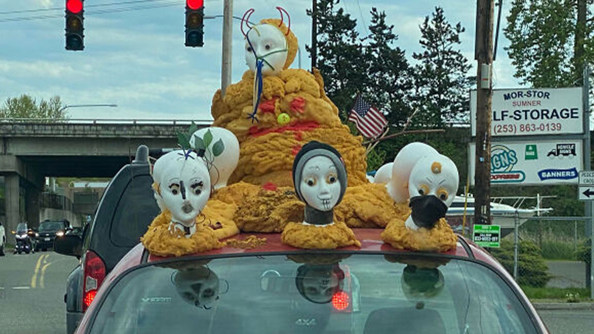 Car with outrageous car mods featuring mannequin heads and unusual foam sculpting on the roof at a red traffic light.