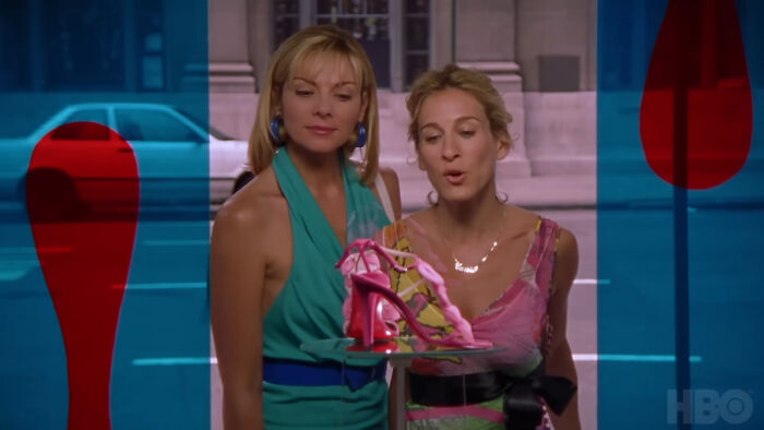 Two women in colorful dresses admiring a pink high-heeled shoe, from a show with hard to love characters.