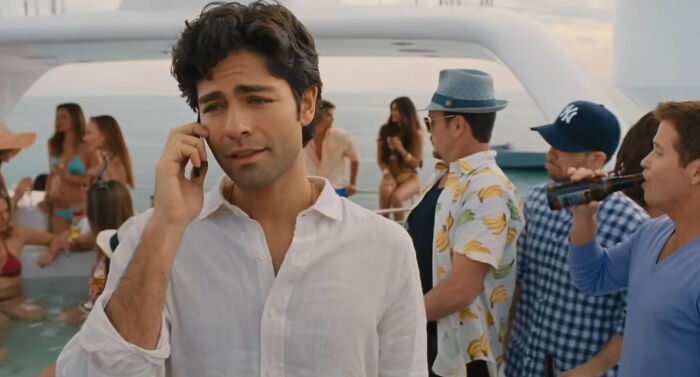 Man in white shirt talking on phone at a party with hard to love TV show characters in the background