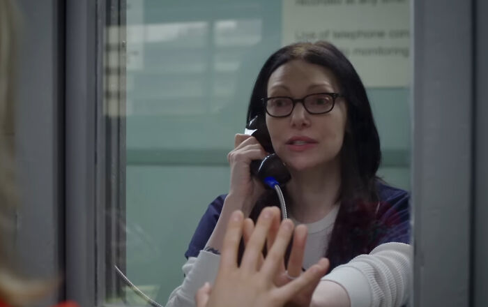 Woman with glasses talking on a phone through a glass partition, depicting characters hard to love in shows.