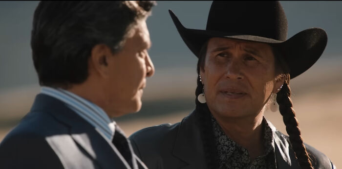 Two men in conversation outdoors, one wearing a black cowboy hat and traditional earrings, showcasing hard to love characters.