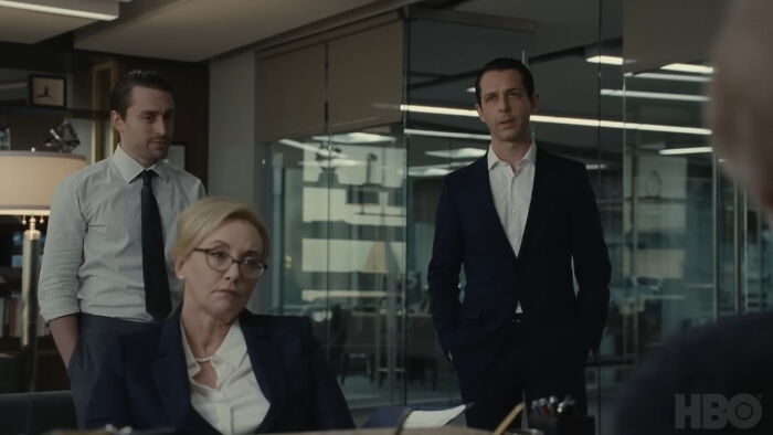 Three serious office workers in formal attire having a tense discussion in a modern glass-walled meeting room.