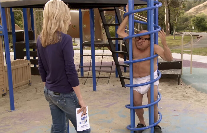 Man in diaper and glasses stuck in playground spiral bars while woman in jeans and blue shirt stands nearby, scene from shows with hard to love characters.