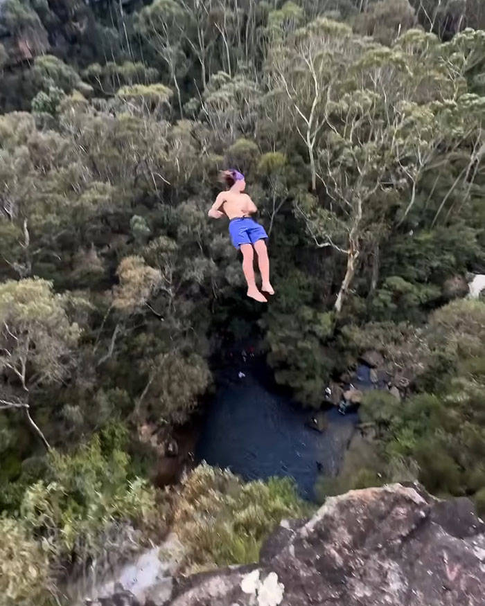 Young daredevil mid-air in shorts diving from high cliff into forested water below during record-breaking dive attempt. Young daredevil mid-air in shorts diving from high cliff into forested water below during record-breaking dive attempt.