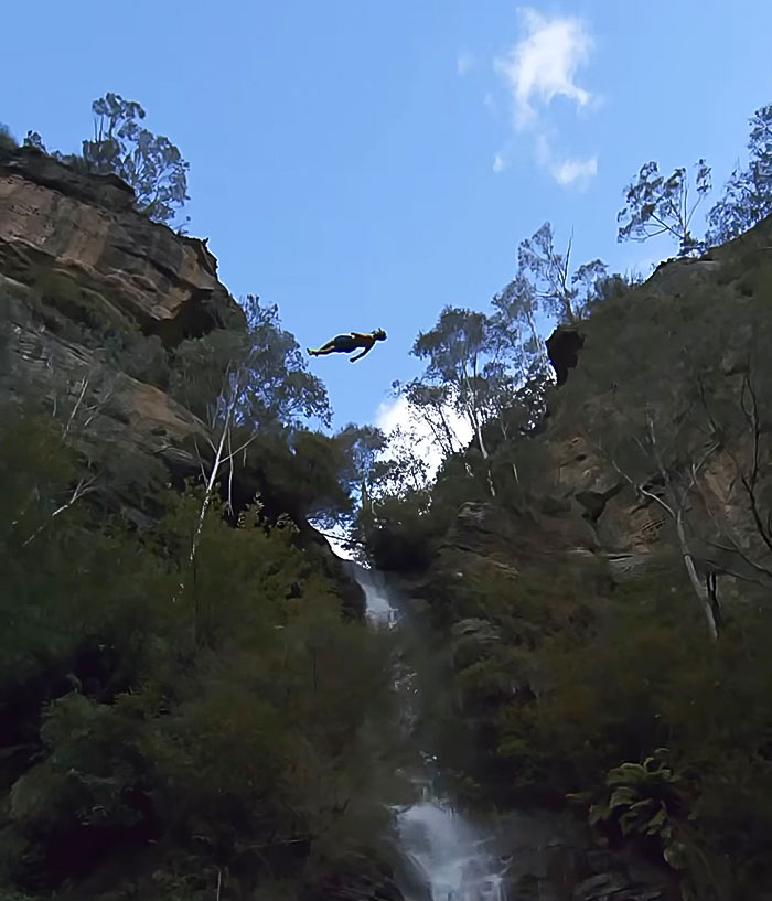21-year-old daredevil mid-air during a record-breaking dive off cliff surrounded by trees and waterfall below. 21-year-old daredevil mid-air during a record-breaking dive off cliff surrounded by trees and waterfall below.