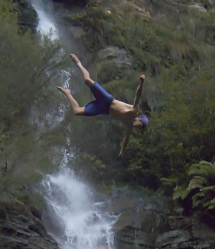 Young daredevil mid-air during a record-breaking dive attempt near a rocky waterfall in a forested area. Young daredevil mid-air during a record-breaking dive attempt near a rocky waterfall in a forested area.