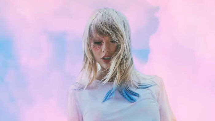 Celebrity with blonde hair and blue streaks wearing a white shirt against a pastel pink and blue sky background.