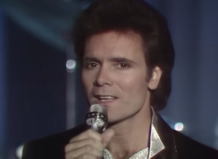 Male celebrity singing into a microphone in a dimly lit setting representing unpopular celebrities many people love but can’t stand