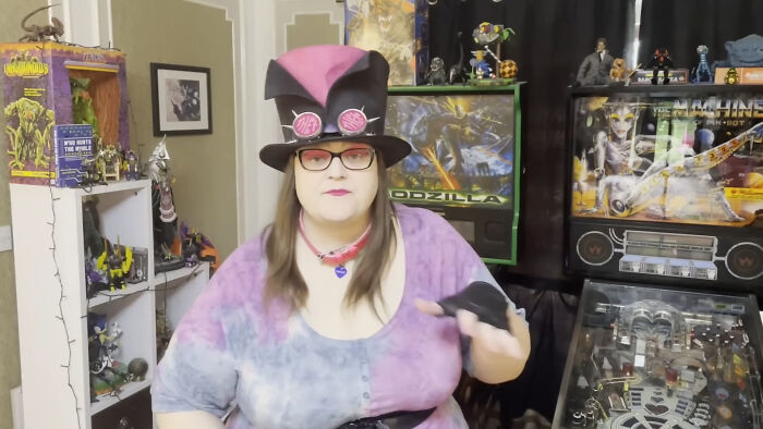 Person wearing a tall hat and glasses, sharing opinions about unsubscribed YouTubers in a room with collectibles and pinball machines.
