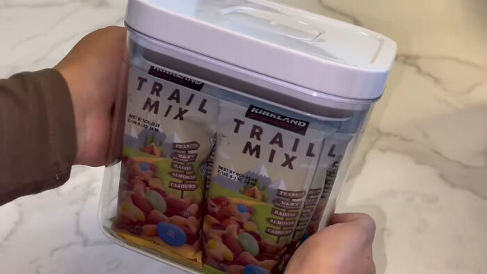 Hands holding a container filled with multiple packs of trail mix, illustrating trends leading to overconsumption.