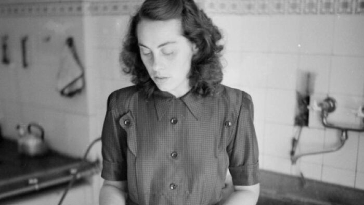 1940s housewife in vintage kitchen setting, wearing a dress and focusing on household chores during wartime era.