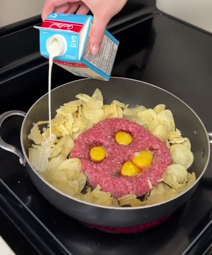 Hand pouring cream into a frying pan with raw ground beef, eggs, and potato chips illustrating overconsumption trends.