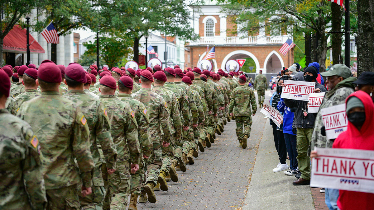 US soldiers in camouflage and maroon berets marching down a street with locals holding thank you signs as a common American scene.