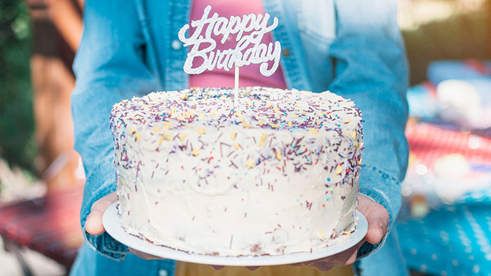 Person in denim jacket holding a birthday cake with sprinkles and a happy birthday topper outdoors in daylight Person in denim jacket holding a birthday cake with sprinkles and a happy birthday topper outdoors in daylight