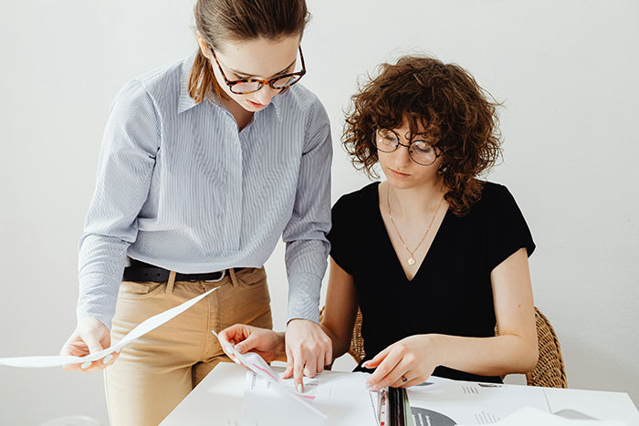 Two women reviewing documents at a desk, the annoyed worker showing frustration with a birthday cake nearby. Two women reviewing documents at a desk, the annoyed worker showing frustration with a birthday cake nearby.