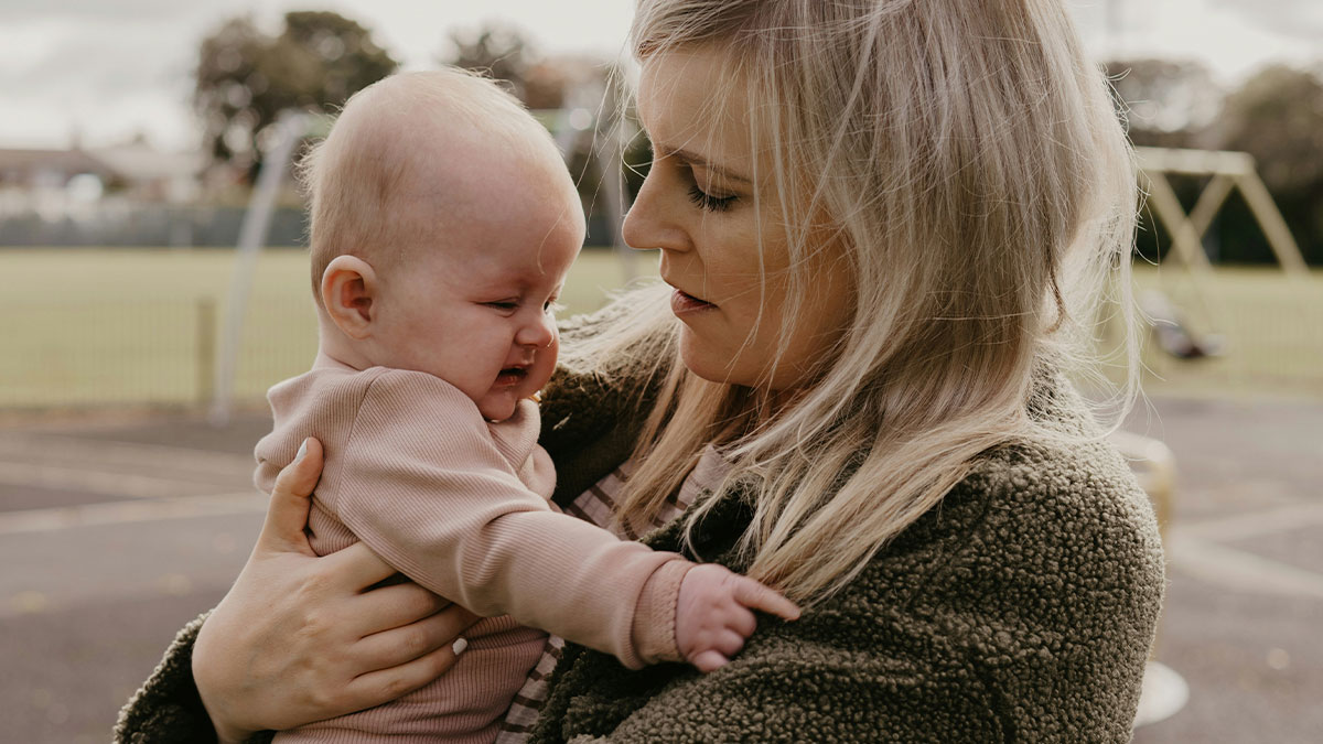 Mother comforting crying 8-month-old twin baby outdoors, illustrating reality check of solo trip while wife cares for children.