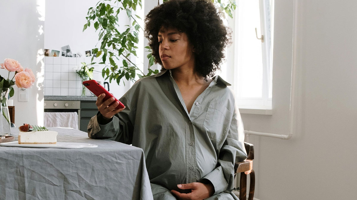 Pregnant lady sitting at table indoors holding phone, concerned about neighbor lurking around her house repeatedly.