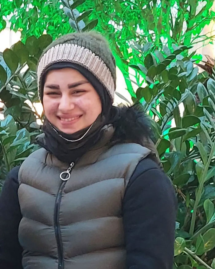 Smiling young woman wearing a winter hat and puffer vest standing outdoors with green leafy plants in the background. Smiling young woman wearing a winter hat and puffer vest standing outdoors with green leafy plants in the background.