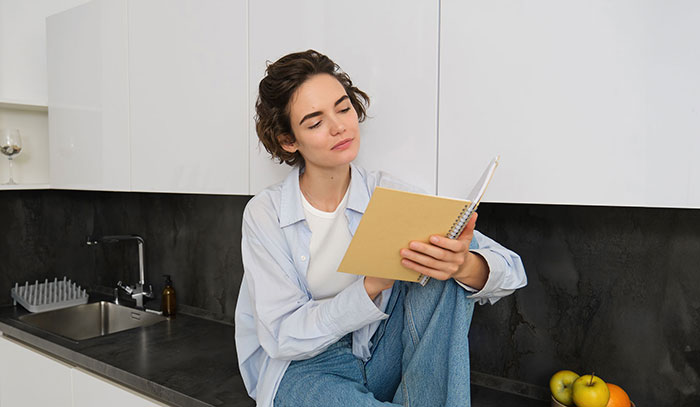 Young woman sitting in kitchen, looking thoughtfully at notebook, depicting emotions related to inheritance and gaslighting issues. Young woman sitting in kitchen, looking thoughtfully at notebook, depicting emotions related to inheritance and gaslighting issues.