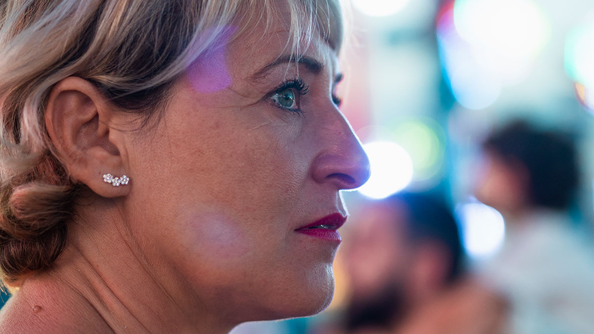 Close-up of woman at concert looking focused as crowd blurs behind her, with purse nearby and ambient colorful lighting.