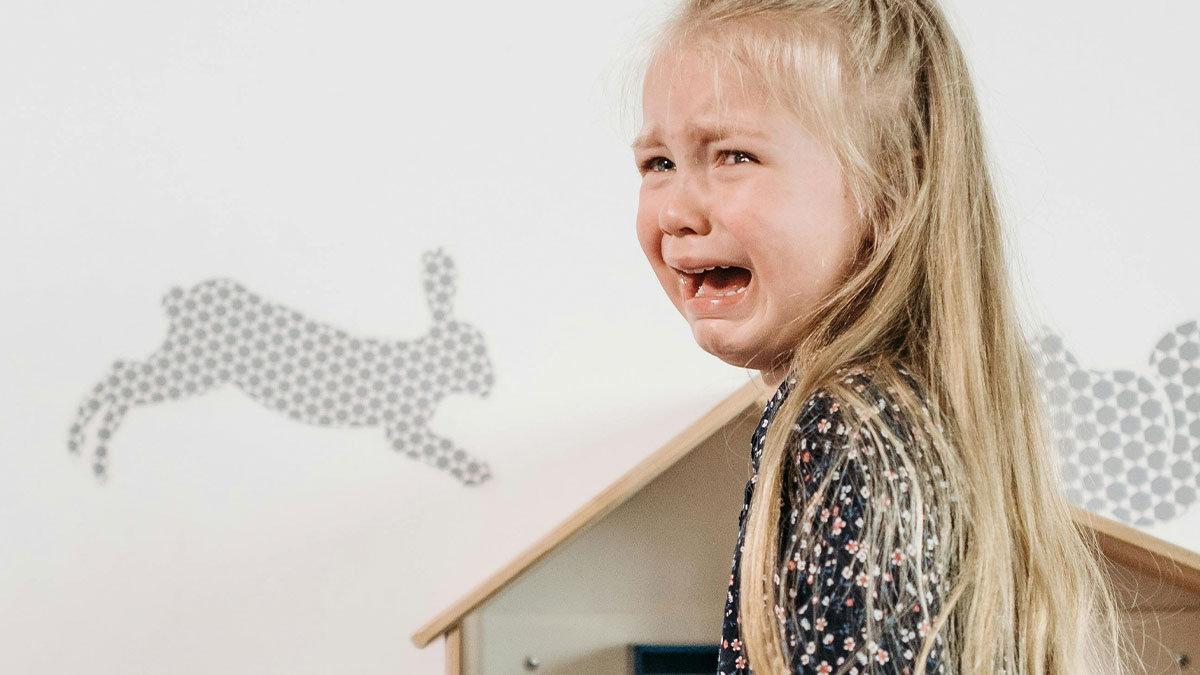 Crying girl with long hair in a patterned dress, reflecting childhood trauma and emotional distress in a playroom setting.