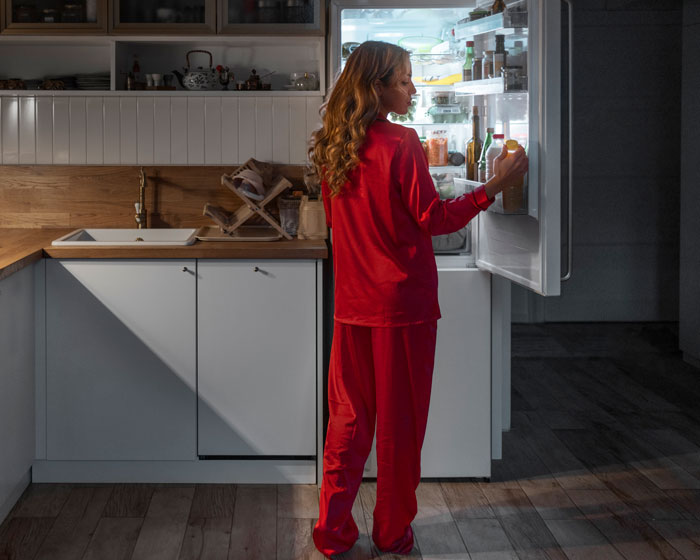 Woman in red pajamas looking inside a fridge at night in a modern kitchen, reflecting quiet living at home.