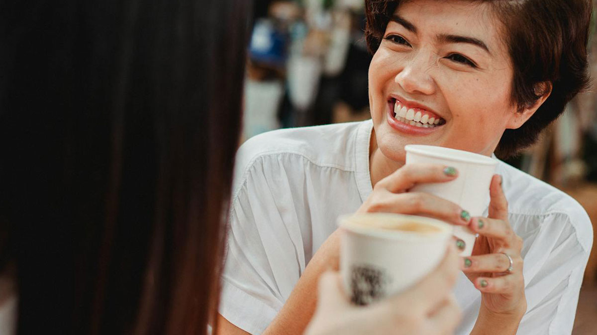 Two women sharing a coffee and smiling while having a casual conversation about disturbing things people overheard.