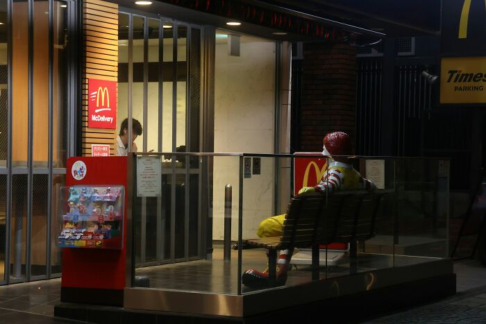 McDonald's employee inside restaurant interacting near entrance with a Ronald McDonald statue seated outside at night.