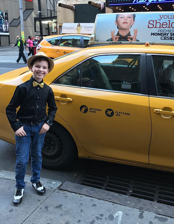Young Sheldon child star in black shirt and bow tie standing next to a yellow taxi in an urban setting. Young Sheldon child star in black shirt and bow tie standing next to a yellow taxi in an urban setting.