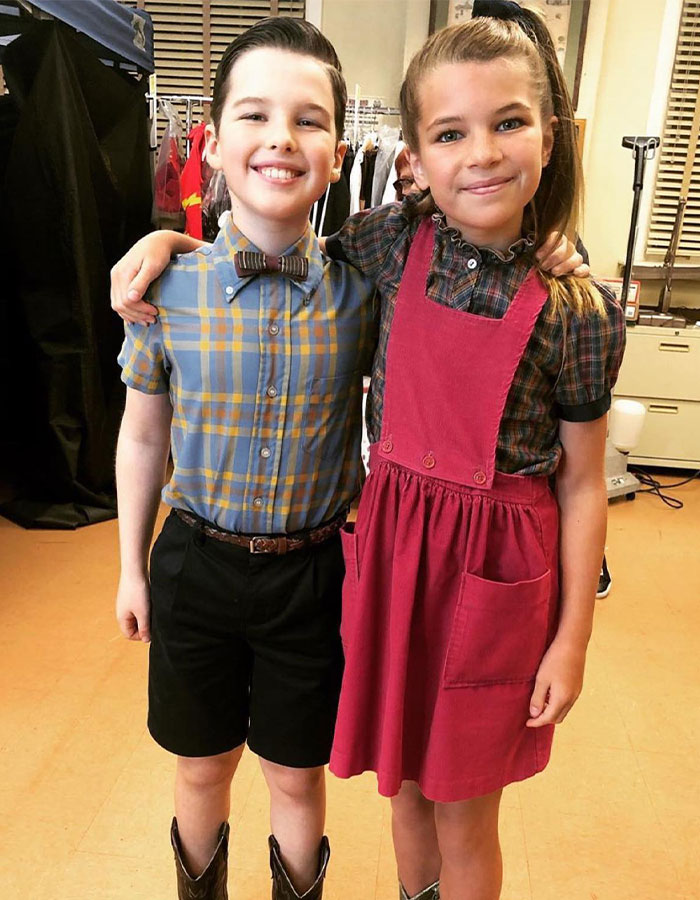 Young Sheldon child star wearing a bow tie and plaid shirt, standing with a girl in a red dress, both smiling. Young Sheldon child star wearing a bow tie and plaid shirt, standing with a girl in a red dress, both smiling.