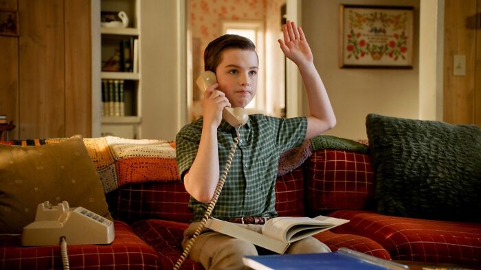 Young Sheldon child star sitting on a couch, holding a phone, and raising his hand in a cozy living room setting. Young Sheldon child star sitting on a couch, holding a phone, and raising his hand in a cozy living room setting.