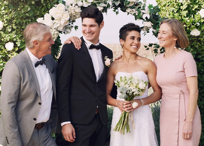 Bride in white dress holding bouquet standing with groom and parents under floral arch, illustrating woman refuses to hand over savings. Bride in white dress holding bouquet standing with groom and parents under floral arch, illustrating woman refuses to hand over savings.