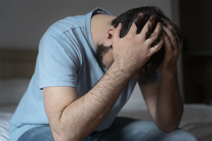 Man sitting on bed holding head in hands, depicting emotional distress after learning about cyber-bullying by wife's family. Man sitting on bed holding head in hands, depicting emotional distress after learning about cyber-bullying by wife's family.