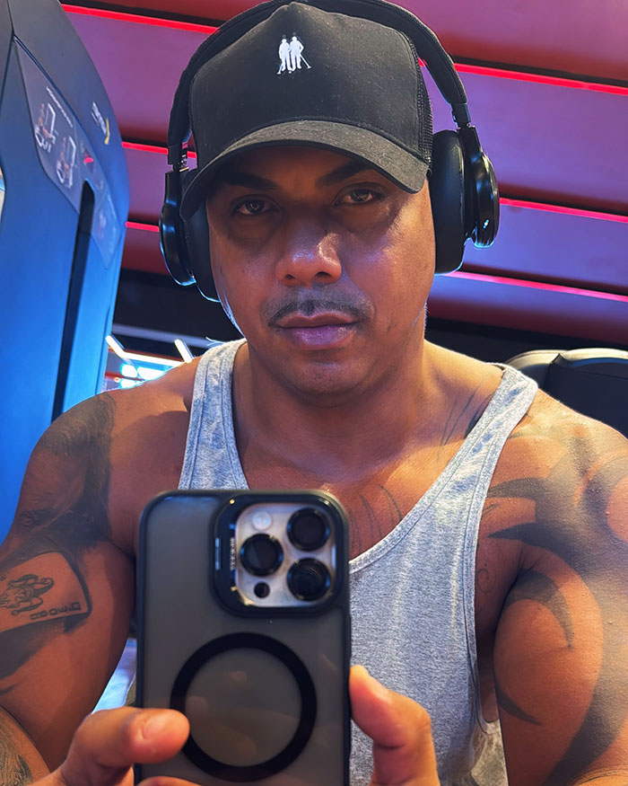Man with headphones and black cap taking a selfie in gym, showcasing muscular arms and tattoo in a sleeveless gray shirt. Man with headphones and black cap taking a selfie in gym, showcasing muscular arms and tattoo in a sleeveless gray shirt.
