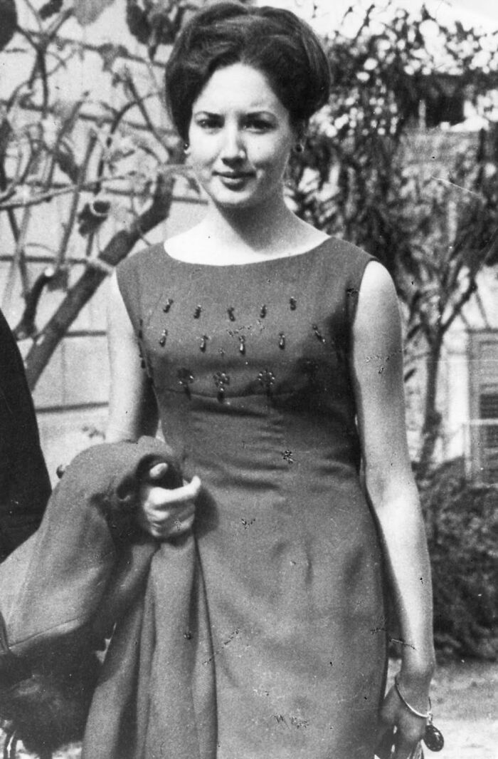 Black and white historical photo of a young woman in a dress holding a coat, with trees and buildings in the background.