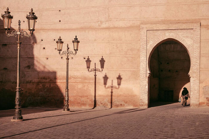 Street lamps casting shadows on an old wall near an arched entrance with a person walking in a candid everyday moment from Asia.