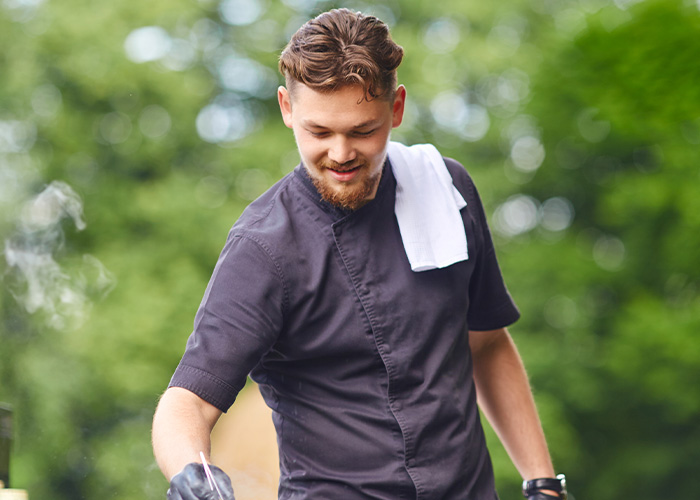 Man wearing a black chef jacket with white towel over shoulder grilling outdoors with smoke and greenery in the background