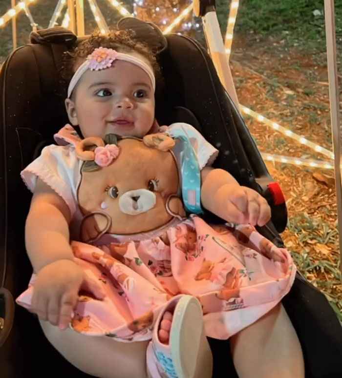 Toddler sitting in car seat wearing pink dress with teddy bear design, surrounded by festive outdoor lights and greenery. Toddler sitting in car seat wearing pink dress with teddy bear design, surrounded by festive outdoor lights and greenery.