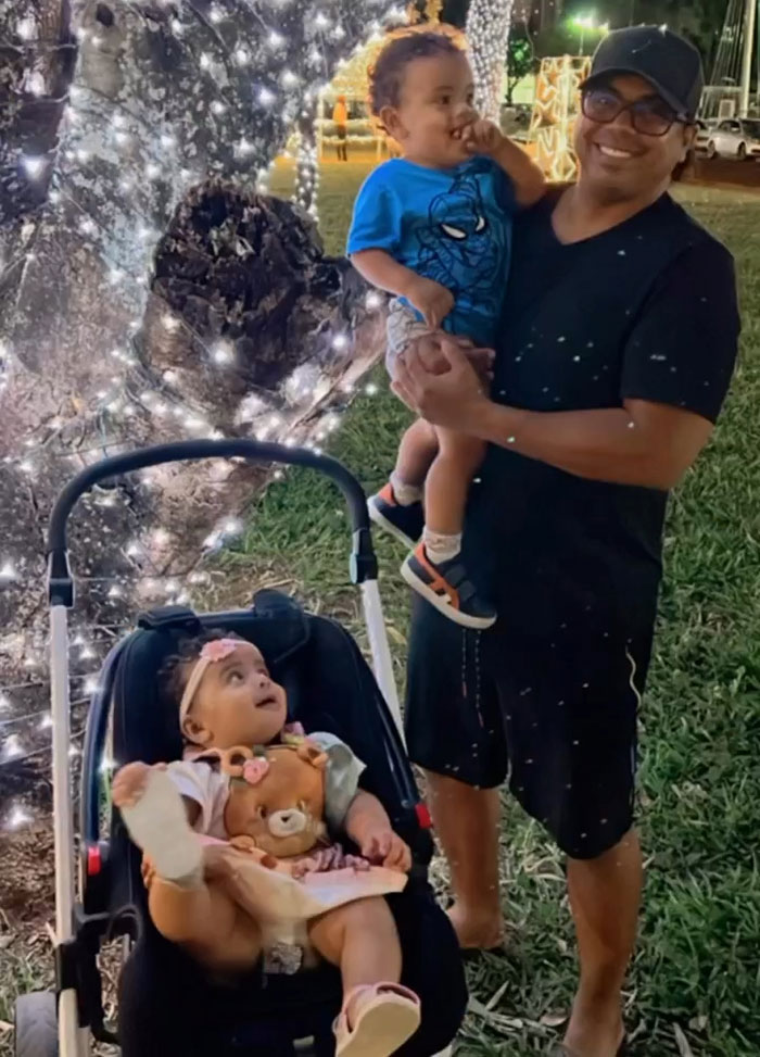 Man holding toddler beside a stroller with a baby and teddy bear near a tree decorated with white lights outdoors at night Man holding toddler beside a stroller with a baby and teddy bear near a tree decorated with white lights outdoors at night