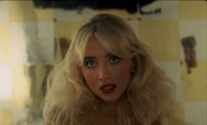 Blonde woman with voluminous hair and red lipstick looks intently in a dimly lit room, related to pole dance video content. Blonde woman with voluminous hair and red lipstick looks intently in a dimly lit room, related to pole dance video content.