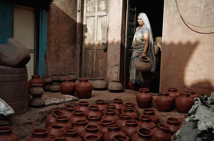 Woman carrying clay pot standing in doorway among rows of earthenware pots in candid everyday moments from Asia.