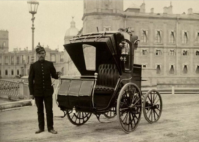 Vintage black early car with large wooden wheels and a man in period uniform standing beside it showing how wild first cars were.