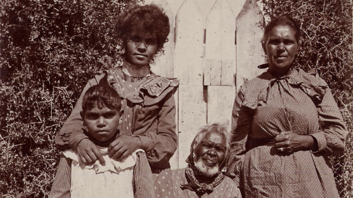 Early 1900s Indigenous Australians posing in traditional and colonial clothing against a wooden fence outdoors.