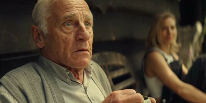 Elderly man looking surprised while holding a cup with a blurred woman in the background in a Netflix movies scene.