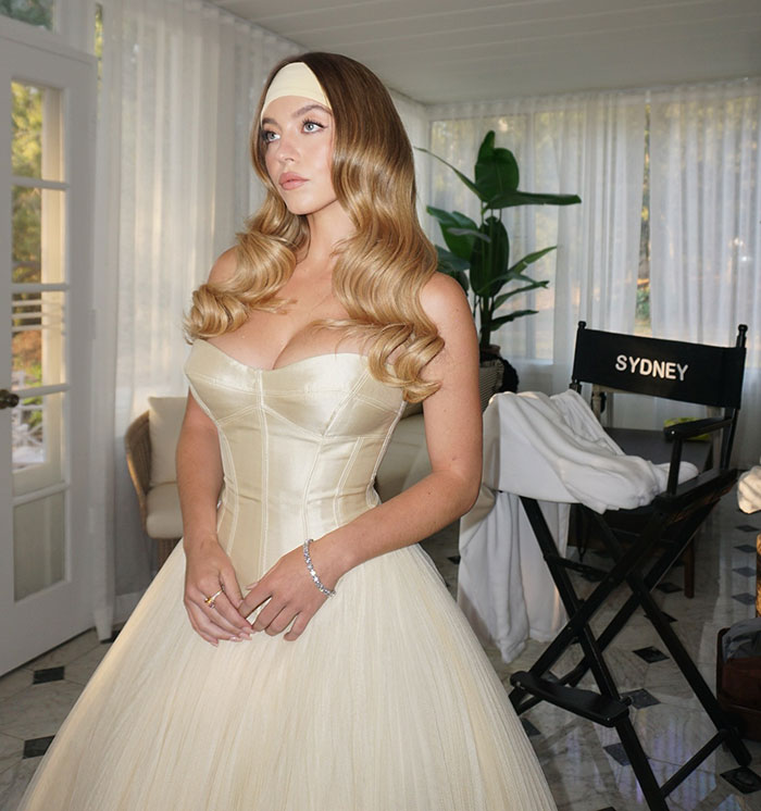 Sydney Sweeney in an elegant cream dress posing indoors near a director’s chair with her name on it. Sydney Sweeney in an elegant cream dress posing indoors near a director’s chair with her name on it.