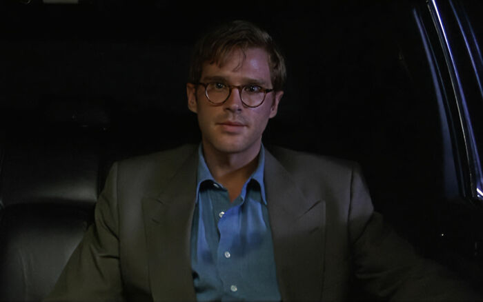 Man wearing glasses and a gray blazer sitting in a car, related to Alicia Silverstone kissing her co-star at age 15.