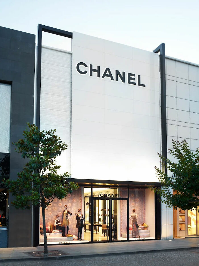 Chanel store exterior with mannequins in window display and trees lining the sidewalk at dusk. Chanel store exterior with mannequins in window display and trees lining the sidewalk at dusk.