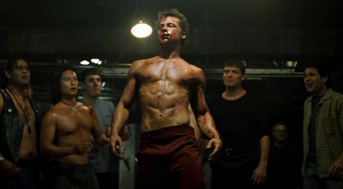 Shirtless young men in a dimly lit room, showcasing wild confidence and boldness among a group of school kids.