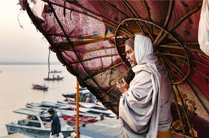 Man wrapped in shawl holding umbrella near docked boats, candid everyday moments from Asia by photographer Gil Kreslavsky
