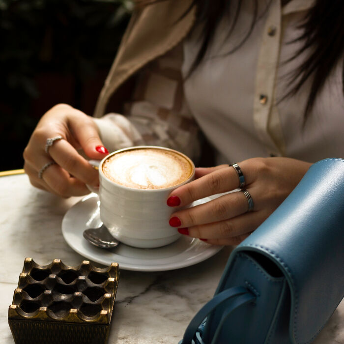 Woman with red nails holding a coffee cup at a marble table next to a blue handbag, illustrating shopping lifestyle.